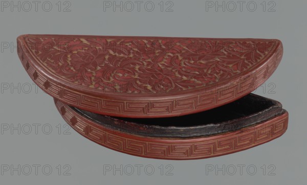 Box with a lid, Chinese art, Chinese culture, Ming 1368–1644, boxes, peonies, meander ornament, leiwen ornament, lemons. Semi-circular box decorated on the lid with a dense pattern composed of peony flowers and leaves. The rim is decorated with a Chinese meander motif. 17th century, China, container. Height 4.4 cm, width 24.0 cm, depth 10.7 cm., A semicircular carved object with intricate floral relief on its top surface and a repeating geometric border along the curved edge, shown partially open to reveal a dark interior. The visible colors include deep red on the carved surfaces, darker reddish-brown along worn areas, black in the interior, and a muted gray background.