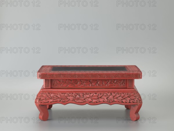 Table-shaped stand, meander and geometric ornament, plant ornament with peonies; Chinese art, Chinese culture; Ming dynasty 1368–1644, 1st half of the 17th century; table; materials/varia; height 12.1 cm, width 26.5 cm, depth 17.8 cm, A small decorative wooden stand painted red with carved floral and leaf patterns and carved geometric borders, featuring a rectangular top with a darker inset panel and supported by curved legs. The visible colors include red, darker red tones, and a dark charcoal or black inset, set against a pale gray background.