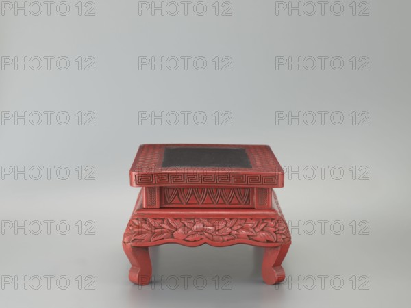 Table-shaped stand, meander and geometric ornaments, plant motifs including peonies; Chinese, Ming dynasty 1368–1644, 1st half of the 17th century; materials/technique: Chinese art; function: table/stand; dimensions: height 12.1 cm, width 26.5 cm, depth 17.8 cm., A small carved wooden pedestal painted red with a slightly darker inset panel on top, featuring a rectangular top surface with geometric border carving, stylized carved patterns on the sides, floral and leaf carvings along the apron, and curved legs; the visible colors are red and a darker muted color on the top panel, set against a pale gray background.