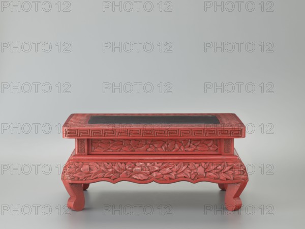 Table-shaped stand, meander and geometric ornament, plant ornament depicting peonies, Chinese art, Chinese culture, Ming 1368–1644, 1st half of the 17th century, table, height 12.1 cm, width 26.5 cm, depth 17.8 cm., A low carved wooden stand painted red with a rectangular dark inset on top, featuring carved floral motifs along the apron and legs, a band of geometric carving beneath the top edge, curved short legs, and a smooth pale gray background; visible colors include red, dark gray, and pale gray.