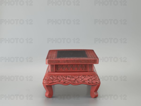 Table-shaped stand, meander ornament, plant ornament, Chinese art, Chinese culture, Ming 1368–1644, peonies, geometric ornament, table, 1st half of the 17th century, China; height 12.1 cm, width 26.5 cm, depth 17.8 cm., A small carved wooden stand painted red with a square top that has a dark inset panel, featuring carved geometric and leaf patterns along the sides and apron, and supported by curved legs; the background is a smooth light gray, and visible colors include red, dark gray-black, and various shades of light gray.
