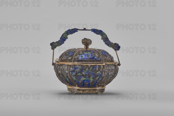 Basket with a lid; maker, Chinese; Canton, China; 19th century; decorated with flowers and ornaments; container; height 9.8 cm, width 10.5 cm, depth 7.5 cm., An ornate metal lidded container with a decorative handle sits against a plain light gray background; the container is bulbous with intricate raised patterns and floral motifs in blue and green enamel set into a tarnished silver or dark metallic body with gold-toned trim and accents, the lid topped by a rounded finial and the hinged handle curved and adorned with blue and green enamel decorations, and the piece rests on a short decorative base with gold-toned elements.