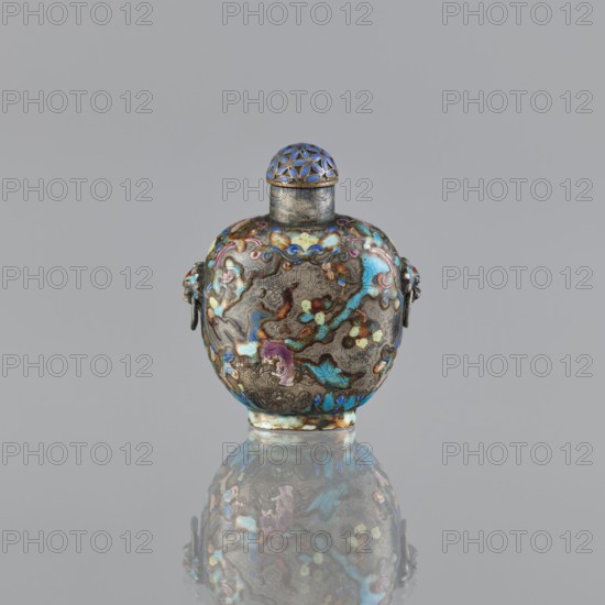Snuffbox with stopper, cloisonné enamel, figural scenes and plant ornament, with handles; maker, Chinese; dated 1929–1930; vessel; height 5.8 cm, width 4.8 cm., A small rounded metal vessel with a short neck and domed lid sits centered against a plain light gray background, the vessel decorated with an ornate, multicolored enamel pattern in shades of blue, turquoise, purple, green, gold, brown, and hints of pink, with symmetrical raised decorative elements on its sides and a patterned lattice on the blue-tinted lid; the surface shows areas of metallic patina and texture, and the object is reflected softly on the glossy surface beneath it, mirroring the same palette and shapes.