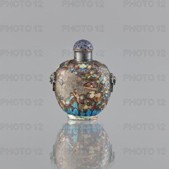 Snuffbox with stopper, cloisonné enamel, figural scenes and plant ornament, handles; maker, Chinese; dated 1929–1930; vessel; height 5.8 cm, width 4.8 cm., An ornate rounded vessel with a domed stopper sits centered against a plain gray background, the vessel displaying a mottled surface of blue, turquoise, green, gold, purple, and brown tones with areas of silver metal showing through; the body has sculpted side elements and subtle raised decorative shapes, and the domed stopper features a geometric openwork pattern in blue and gold; the vessel and its colorful surface are reflected below on a glossy surface, showing a slightly blurred mirror image.