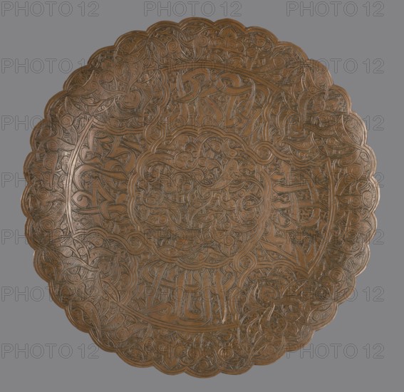 Plate, Islamic art, Arabic culture, geometric motifs, arabesque ornament, Arabic inscriptions, Arabic language. Flat plate without a distinct rim. Decorated with a dense arabesque in the hataji style and an Arabic inscription in suius. Early 20th century, Egypt, Syria . Vessel; height 1.6 cm, diameter 29.6 cm., A round, scalloped-edge metal plate with an intricate raised pattern covering its surface, featuring swirling, interlaced lines and botanical motifs arranged concentrically; the plate has a warm brown, coppery tone with darker engraved lines and subtle highlights, set against a neutral gray background.