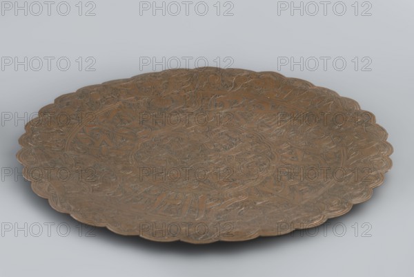 Plate, flat without a distinct rim. Decorated with a dense arabesque in the hataji style and an Arabic inscription in suius. Early 20th century. Islamic art; Arabic language; geometric motifs; arabesque ornament; Arabic inscriptions., Possibly Egypt or Syria. Vessel; height 1.6 cm; diameter 29.6 cm., A shallow round metal tray with a scalloped rim fills most of the image, showing intricate incised patterns across its surface including spirals, interlacing shapes, and floral-like motifs; the tray appears in shades of brown and bronze with darker engraved lines adding contrast, and it rests on a smooth pale gray background that creates a soft shadow beneath the tray.