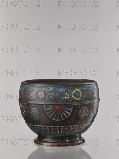 Flowerpot, Shimasa creator/workshop role uncertain, Japanese, Meiji period late 19th century, 1868–1912. Champlevé enamel on bronze jardinière with floral ornament. Vessel; height 30.4 cm, diameter 41.2 cm. Unknown provenance., The image shows a decorative rounded vessel with a short pedestal base, featuring ornate floral and geometric designs in a palette of blue, red, yellow, green, and dark brown, with accents of gold and black; the top band displays a series of stylized flowers on a blue background, the midsection has larger fan-shaped and circular floral motifs with red and blue petals on a dark brown ground, and the lower band contains repeating arch-like patterns in blue and red; the vessel sits on a reflective surface against a neutral pale gray background.