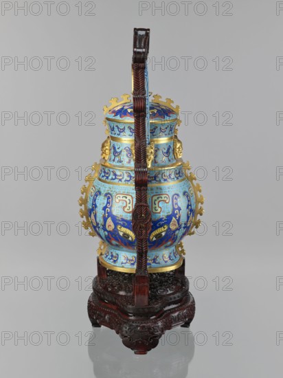 You-type vessel with a lid on a stand, maker, Chinese, Qing dynasty, Qianlong period 1736–1795. Cloisonné cellular enamel masterpiece; ritual/ceremonial you vessel originally used for storing alcoholic beverages form dates to Shang dynasty. Elaborate, precise, multicolored cloisonné decoration on body and lid depicting a taotie mask and stylized birds—motifs borrowed from archaic bronzes. Plastic insets on the sides and animal-head terminals on both sides of the movable handle. Wooden base mattly polished rosewood elongated with bas-relief decoration: symbolic bats and spiral ornament. Materials: cloisonné enamel, wood stand. Vessel dimensions: height 39.0 cm, width 24.0 cm, depth 19.0 cm, weight 5.6 kg. Stand dimensions: height 55.0 cm, width 30.0 cm, depth 18.0 cm, weight 1.8 kg., An ornately decorated vessel sits on an intricately carved wooden stand with a tall carved wooden handle arching over it; the vessel is predominantly shades of blue with accents of gold and turquoise, featu...