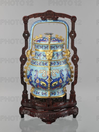 You-type vessel with a lid on a stand, cloisonné enamel, Qianlong period 1736–1795, Qing dynasty, China. Elaborate, precise, multi-colored cellular enamel decoration on body and lid depicting a taotie mask and stylized bird images, motifs borrowed from archaic bronzes. Plastic insets decorate the sides and animal heads are placed on both sides of the movable handle. The you vessel with a lid and movable handle was originally used for storing alcoholic beverages during the Shang Dynasty. Wooden stand mattly polished rosewood, elongated shape, decorated with bas-reliefs of symbolic representations bats and spiral ornament. Height vessel 39.0 cm; width 24.0 cm; depth 19.0 cm; weight 5.6 kg. Stand: height 55.0 cm; width 30.0 cm; depth 18.0 cm; weight 1.8 kg. Chinese art; ritual/ceremonial vessel; masterpiece., An ornately decorated vessel sits within a carved wooden stand and frame; the vessel is domed with a lid and a rounded body and features an overall palette of bright blue and light b...