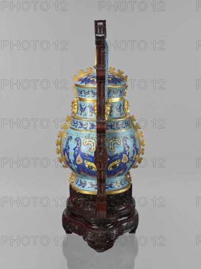 You-type vessel with a lid on a stand, Chinese art, Qing dynasty 1644–1911, Qianlong period 1736–1795; cloisonné cellular enamel ritual/ceremonial vessel you decorated with images of birds and a taotie mask in powerful stylization; decorative motifs and plastic insets borrowed from archaic bronzes, with animal-head terminals on the movable handle; wooden elongated stand mattly polished rosewood decorated with bas-relief bats and spiral ornament; originally used as a vessel for storing alcoholic beverages during the Shang Dynasty. Dimensions — vessel: H 39.0 cm, W 24.0 cm, D 19.0 cm, weight 5.6 kg; stand: H 55.0 cm, W 30.0 cm, D 18.0 cm, weight 1.8 kg., A decorative vessel with a rounded body and a domed lid sits on an ornate carved wooden stand, the vessel suspended by a vertical carved wooden handle that rises above it; the vessel is richly patterned in bright blue and light blue with accents of gold and touches of red and green, and features symmetrical decorative motifs and gold orn...