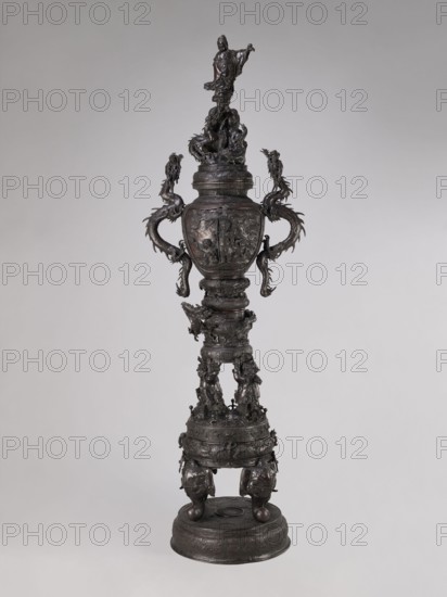 Incense burner koro, Meiji period late 19th century, Japan. Bronze vessel incense burner with dragon-shaped handles; iconography includes dragons, devils, elephants, zodiac signs, peacocks, pine trees, peonies, bodhisattva, and Amaterasu. Height 255.0 cm; width 78.0 cm; depth 46.0 cm., A tall ornate metal urn-like sculpture stands against a plain light-gray background, rendered in dark brown and black tones with subtle lighter highlights; it features a sculpted figure at the top standing on a textured base, flowing drapery, and multiple dragon-like serpentine forms emerging as handles that curve outward and downward; the central body is rounded with detailed relief scenes and decorative bands, leading to an elongated segmented stem adorned with carved figures and ornamental motifs; the lower portion expands into a drum-like base covered in embossed patterns and supported by bulbous feet with animal-head elements, all exhibiting a metallic sheen and intricate surface texture.