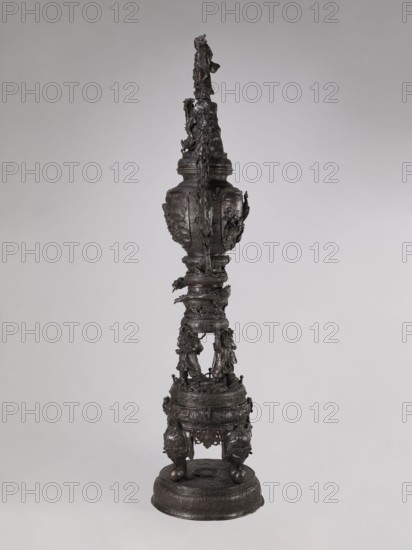 Incense burner koro, Meiji period late 19th century, Japan. Bronze vessel incense burner with dragon-shaped handles; decorated with dragons, devils, elephants, zodiac signs, peacocks, pine trees, peonies, bodhisattva imagery, and Amaterasu mythology. Function: incense burner. Dimensions: height 255.0 cm, width 78.0 cm, depth 46.0 cm., A tall dark bronze sculptural object stands against a pale gray background, featuring a layered vertical form with ornamental textures and relief work; the piece has a rounded base rising to a circular plinth with carved motifs, supported by rounded feet, above which a drum-like section with elaborate surface decoration and a group of small humanlike figures in dynamic poses connects to a slender central column wrapped with sculpted vine- or rope-like elements; higher up there is a vase-shaped midsection with raised decorative panels and more relief ornamentation, topped by a tapering upper shaft that culminates in a small standing figure; the overall col...