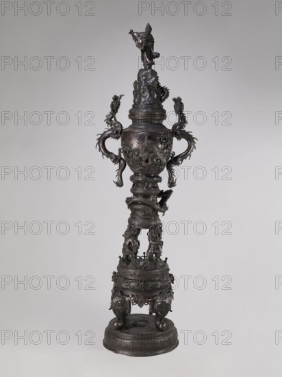 Incense burner koro, Meiji period late 19th century, Japan; bronze vessel/incense burner with dragon-shaped handles; decorated with dragons, devils, elephants, zodiac signs, peacocks, pine trees, peonies, bodhisattva imagery, and Amaterasu; Japanese art; height 255.0 cm, width 78.0 cm, depth 46.0 cm, A tall, dark metallic sculptural object stands against a neutral light gray background, with a shiny blackish-brown patina and areas of darker and slightly reddish-brown hues. The piece has an elongated vertical form composed of several stacked sections: a rounded ornate base with a circular platform, a midsection supported by sculpted animal-like legs and rounded feet, and a narrower column that rises to a bulbous vessel-like portion. The vessel section features two large ornate handles shaped like curling dragon or serpentine figures extending outward and upward, and the surface of the vessel and adjoining sections is richly textured with raised decorative motifs and sculpted figures. Hi...