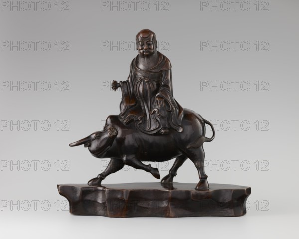 Laozi on a Buffalo, Chinese, Qing dynasty 1644–1911, 19th century; sculpture/figurine depicting Laozi ca. 550–500 BC riding a buffalo; materials not specified; dimensions: height 39.5 cm, width 41.0 cm, depth 16.5 cm; subjects: animals, buffaloes., A dark brown wooden sculpture shows a robed bearded man seated on the back of a water buffalo, both resting on a carved base that is also dark brown; the man wears flowing layered robes with detailed folds and has a calm facial expression, a long beard, and a rounded head, while the buffalo has a curved horn, visible ears, a short tail, and a muscular body with one front leg lifted; the surface has polished highlights and subtle variations of brown and black tones against a neutral gray background.