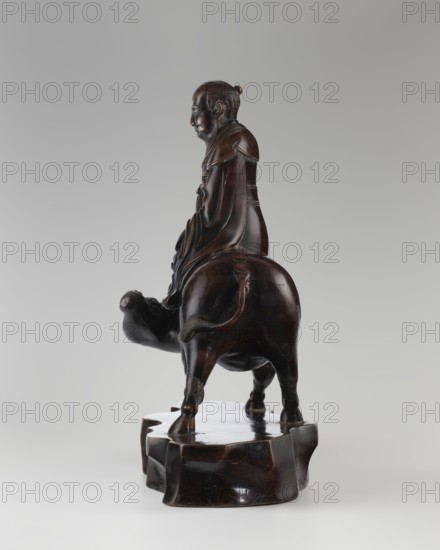 Laozi on a Buffalo, Chinese, Qing dynasty 1644–1911, 19th century. Sculpture/figurine depicting Laozi ca. 550–500 BCE riding a buffalo. Materials: Chinese art; dimensions: height 39.5 cm, width 41.0 cm, depth 16.5 cm. Subjects: animals, buffaloes., A dark brown carved figure seated on the back of a stout animal atop a carved base is shown against a light gray background; the figure has a rounded head with a small topknot and a detailed facial profile, dressed in flowing robes with visible folds and layered shoulder pieces, and the animal beneath has a rounded body, short legs, a curved tail, and a slightly raised head; the base is irregularly shaped and shares the same dark brown color, with smooth polished surfaces and subtle variations in sheen.