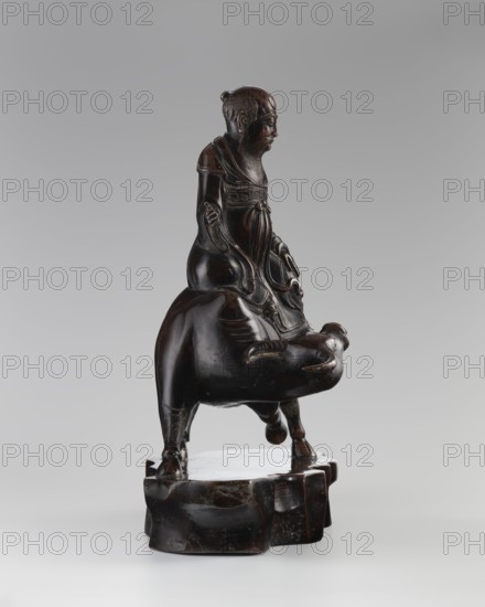 Laozi on a Buffalo, Chinese, Qing dynasty 1644–1911, 19th century. Sculpture/figurine depicting Laozi ca. 550–500 BCE seated on a buffalo; materials not specified. Dimensions: height 39.5 cm, width 41.0 cm, depth 16.5 cm. Subjects: animals, buffaloes., A dark brown sculptural figure made of a glossy material sits on a rounded irregular base of the same color, showing a seated humanlike figure with a long beard and hair tied back, wearing layered flowing garments with carved folds and a tied sash, positioned atop an animal with a rounded body and short legs; the surface displays highlights and subtle variations of darker and lighter brown tones against a neutral gray background.