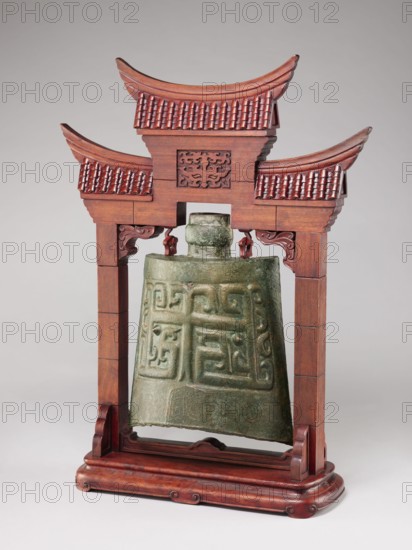 Bell on a stand in the shape of a paifang gate., Chinese, Qing dynasty 1644–1911. Bronze zhong-type bell 18th century, missing the upper part of the shaft used for hanging the part above the ring, decorated with an archaized pattern resembling a taotie mask. Wooden stand, gilded, in the shape of a paifang gate with a mount for hanging the bell; after conservation the stand consists of three elements 19th century. Categories: bells, bronzes, taotie iconography, gates, musical instruments, percussion instruments, idiophones. Dimensions: stand height 64.5 cm; bell height 34 cm., A decorative hanging bell suspended in a carved wooden frame shaped like a small pagoda is shown, with the bell made of greenish tarnished metal featuring raised geometric and curvilinear relief patterns, and the wooden frame finished in a reddish-brown tone with carved roof tiles, curved eaves, ornamental brackets, and a rounded base.