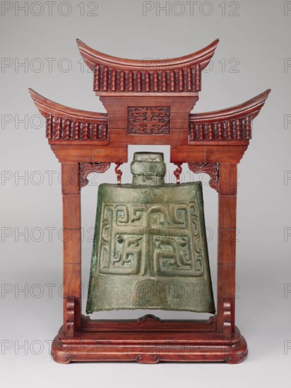 Bell on a stand in the shape of a paifang gate. Chinese; Qing 1644–1911. Bronze zhong-type bell 18th century, missing the upper part of the shaft used for hanging the part above the ring, decorated with an archaized pattern resembling a taotie mask. Wooden stand, gilded, in the shape of a paifang gate with a mount for hanging the bell; after conservation the stand consists of three elements 19th century. Subjects/attributes: bells, bronzes, taotie iconography, gates, musical percussion idiophone. Dimensions: stand height 64.5 cm; bell height 34 cm., A greenish bronze bell with a textured patina and raised geometric relief patterns hangs from a reddish-brown wooden frame carved with ornate roof-like curves and tiled details; the frame has a polished finish with carved brackets and curved base elements, and the background is a neutral light gray.