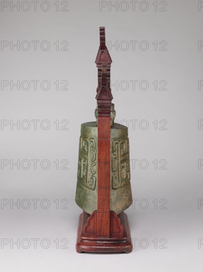 Bell on a wooden stand in the shape of a paifang gate, gilded mount for hanging the bell. Bronze zhong-type bell 18th century, missing the upper part of the shaft used for hanging, decorated with an archaized pattern resembling a taotie mask. Stand 19th century consists of three elements after conservation. Chinese; Qing dynasty 1644–1911. Materials: bronze, wood. Categories: bells, bronzes, taotie iconography, gates, musical/percussion idiophones. Dimensions: stand height 64.5 cm; bell height 34 cm., The image shows an object consisting of a greenish bronze bell with a weathered surface and carved raised patterns hanging within a reddish-brown wooden stand; the stand has a vertical central post with decorative stepped and triangular elements at the top and a carved base with curved details, all set against a neutral light gray background.