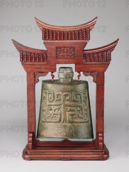 Bell on a stand in the shape of a paifang gate., Chinese art; Chinese culture; Qing 1644–1911. Zhong-type bronze bell decorated with an archaized tao-tie–like mask; missing the upper part of the shaft used for hanging the part above the ring. Wooden gilded stand in the shape of a paifang gate with a mount for hanging the bell; after conservation the stand consists of three elements. Bell 18th century; stand 19th century; China. Dimensions: stand height 64.5 cm; bell height 34 cm., A carved wooden stand shaped like a small ornate gateway supports a hanging bell; the stand is a rich reddish-brown with curved eaves, decorative tiled roof patterns, scrollwork brackets, and a carved panel in its upper center, and it rests on a rounded stepped base of the same color. The bell suspended within is greenish with areas of brown and gray patina, showing raised abstract geometric relief patterns and a rounded top mount. The background is a smooth light gray.