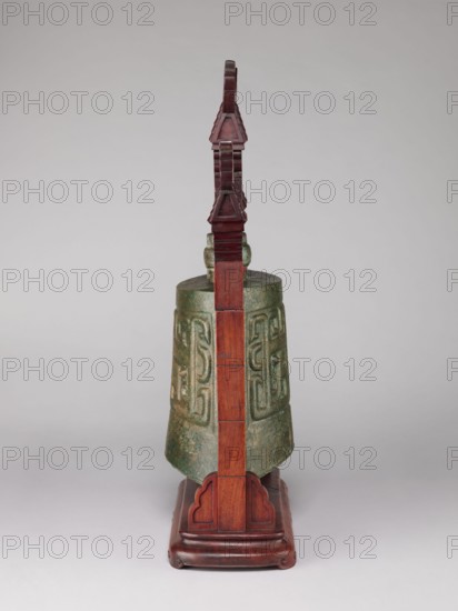 Bell on a stand in the shape of a paifang gate., Chinese, Qing dynasty 1644–1911. Zhong-type bronze bell, missing the upper part of the shaft used for hanging the part above the ring, decorated with an archaized pattern resembling a taotie mask. Wooden stand, gilded, in the shape of a paifang gate with a mount for hanging the bell; after conservation the stand consists of three elements. 18th century bell, 19th century stand. Bell height 34 cm; stand height 64.5 cm. Materials: bronze, wood, gilding. Genres: bells, bronzes, taotie iconography, gates, musical/percussion idiophones., A vertical bronze bell with a greenish patina hangs from a tall reddish-brown wooden stand; the bell shows raised decorative patterns and a weathered surface, and the stand has a carved base and a stepped, ornamental top, all set against a neutral light gray background.