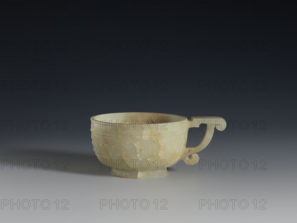 Bowl, Chinese, Qing dynasty 1644–1911, 18th century, China; porcelain vessel with rueding pattern and knobs ornament, gui vessel form; height 3.8 cm, width 9.6 cm, A pale yellowish-green carved cup with a rounded body and a decorative handle sits on a smooth gray surface against a dark gray to near-black gradient background; the cup shows a mottled surface with areas of creamy beige and light brown veining, a band of small raised circular bumps around its exterior, a slightly flared rim, and a short circular foot, and its handle has a curved, scroll-like end.