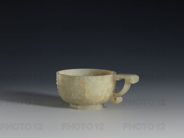Bowl, Chinese, Qing dynasty 1644–1911, 18th century. Porcelain vessel with ruding pattern and knobs ornament, gui vessel form. Height 3.8 cm; width 9.6 cm., A small shallow cup with a single curved handle sits on a flat surface, carved from a pale, translucent stone showing creamy white, light beige, and faint yellowish tones with subtle cloudy variations; the cup has a rounded body on a short foot, a slightly flared rim, a decorative band of raised round dots around its exterior, and a handle with a curled end, all shown against a smooth gradient background shifting from dark charcoal at the top to lighter gray near the bottom and casting a soft shadow to one side.