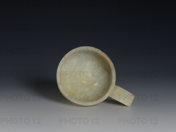 Bowl, Qing dynasty 18th century, 1644–1911, China; ceramic vessel with ruding pattern and knobs ornament, gui vessel form; height 3.8 cm, width 9.6 cm., A round shallow bowl-like object with a short handle, made of a smooth translucent stone showing pale green, creamy white, and light amber tones with cloudy veining; it sits on a flat surface casting a soft shadow and is photographed against a dark gradient background that transitions from deep charcoal at the top to medium gray toward the bottom.