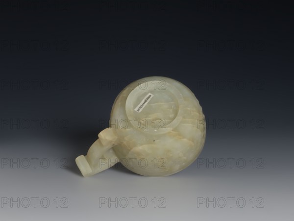 Bowl, Chinese, Qing dynasty 1644–1911, 18th century. Porcelain vessel with ruding pattern, gui vessel form, knobs ornament. Height 3.8 cm, width 9.6 cm., A round, pale greenish-white jade object with a short curved handle rests on a smooth gray surface against a dark gradient background; the jade shows translucent areas and cloudy patches of cream and light yellow, with subtle striations and polished sheen, and a few small darker inclusions are visible in the stone.