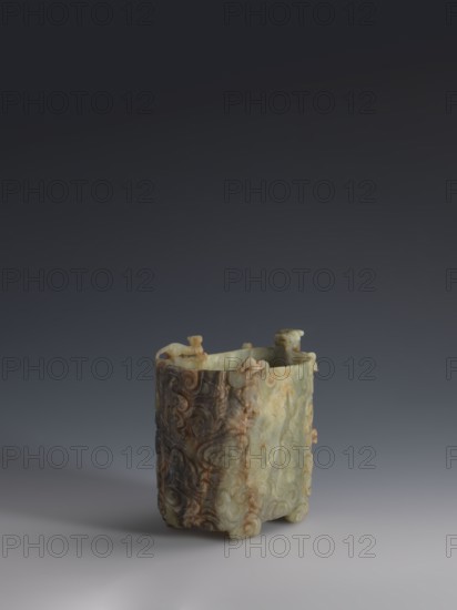 Vessel., Chinese, Qing dynasty 1644–1911, early 18th century. Gray-green jade, hollowed; bas-relief motifs of clouds and mythological creatures covering the entire surface. Tall vessel on four feet with rounded edges; dragon-shaped handle on the narrower side. Height 9.0 cm, width 14.0 cm, depth 6.5 cm. Icons: dragons, clouds., An upright rectangular carved object with rounded corners resting on a smooth surface, showing a mix of pale green, creamy beige, brown, and darker brown tones; the surface is covered in swirling carved motifs and raised decorative elements, with small sculpted protrusions along the upper rim and short rounded feet at the base; the background is a smooth gradient from dark gray at the top to lighter gray near the bottom.