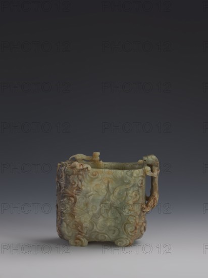 Vessel, Chinese, Qing dynasty 1644–1911. Early 18th century. Gray-green jade, hollowed. Tall vessel on four feet with rounded edges; dragon-shaped handle on the narrower side. Entire surface covered in bas-relief motifs of clouds and mythological creatures dragons. Height 9.0 cm; width 14.0 cm; depth 6.5 cm., An ornate carved vessel sits centered against a dark gray to black background, made of a greenish stone with areas of brown and tan discoloration, featuring a square body with rounded feet and flowing raised scroll and cloud motifs across its surfaces, a carved loop handle on one side with organic knob-like elements at the rim, and the stone showing polished smooth areas and rougher carved reliefs in shades of pale green, olive, brown, and beige.
