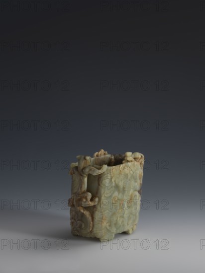 Vessel., Chinese, Qing dynasty 1644–1911, early 18th century. Gray-green jade, hollowed; bas-relief motifs of clouds and mythological creatures covering the entire surface. Tall vessel on four feet with rounded edges; dragon-shaped handle on the narrower side. Height 9.0 cm, width 14.0 cm, depth 6.5 cm. Icons: dragons, clouds., The image shows a carved object with a pale green and light brown color palette sitting on a smooth surface against a dark gradient background; the object has an ornate surface with swirling carved patterns and rounded protrusions, an open top revealing a hollow interior, curved handles or decorative extensions on its sides, and a slightly irregular rectangular shape with carved feet at the base.
