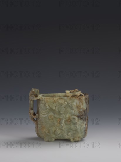 Vessel., Chinese; Qing dynasty 1644–1911. Early 18th century. Gray-green jade, hollowed; bas-relief motifs of clouds and mythological creatures covering the entire surface. Tall vessel on four feet with rounded edges; dragon-shaped handle on the narrower side. Height 9.0 cm; width 14.0 cm; depth 6.5 cm. Icons: dragons, clouds., A small carved object with a square form sits centered against a gradient background that transitions from dark gray at the top to light gray at the bottom; the object is predominantly pale green with areas of brown and darker green, and it features intricate raised swirling patterns and carved details on its surfaces, including a handle on one side with looping shapes and carved decorative elements along the top and corners, and subtle shadows beneath it on the light gray surface.