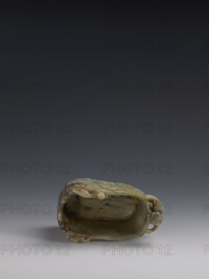 Vessel., Chinese, Qing 1644–1911. Early 18th century. Gray-green jade, hollowed. Tall vessel on four feet with rounded edges; dragon-shaped handle at the narrower side. Entire surface covered in bas-relief motifs of clouds and mythological creatures dragons, clouds. Height 9.0 cm; width 14.0 cm; depth 6.5 cm., A carved object rests on a smooth surface, shown from a low frontal angle with a backdrop that shifts from dark gray at the top to lighter gray near the bottom. The object has an elongated hollow form with an irregular opening and textured exterior, displaying a mix of green, pale green, brown, and beige tones with mottled patches and subtle veining. The surface includes carved protrusions and rounded shapes around the rim and one side, and the interior shows smoother, slightly glossy surfaces with similar green and brown coloration. Shadows fall beneath and slightly to one side of the object, and the overall scene is evenly lit with soft light.