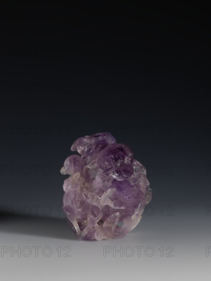 Pomegranate-shaped vessel, Chinese, Qing dynasty 1644–1911, 18th–19th century. Portrays pomegranates and frogs; vessel. Height 10.2 cm, width 9.6 cm, diameter 7.2 cm., A polished translucent purple stone with areas of lighter lavender and clear sections sits on a smooth gray surface, casting a soft shadow; the stone has an irregular, rounded shape with carved or naturally formed flowing contours and layered, petal-like protrusions, and the background fades from dark charcoal at the top to mid-gray behind the stone.