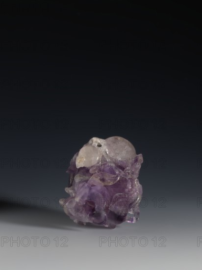 Pomegranate-shaped vessel, Qing dynasty 1644–1911, 18th–19th century, China; porcelain vessel decorated with pomegranates and frogs; height 10.2 cm, width 9.6 cm, diameter 7.2 cm., An irregularly shaped translucent object sits on a smooth surface against a dark gradient background, showing a mix of purple and pale lavender hues with areas of clear, glassy transparency and some cloudy inclusions; the form features rounded bulbous sections and flowing, carved-like contours with glossy highlights and subtle shadows, and the surface reflects light creating small bright spots and softer gleams in tones of purple, clear, and pale gray.