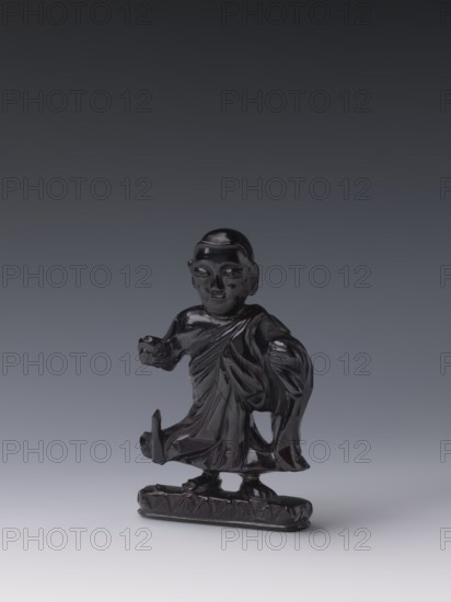 Buddhist monk, Chinese, Qing style, Chinese art, Chinese Buddhism, Qing 1644–1911, Luohan iconography, 19th century, China, sculpture figurine, height 11.8 cm, width 7.1 cm, depth 2.0 cm, A glossy dark figurine of a robed figure stands on a small rectangular base, the surface reflecting light and appearing black with subtle highlights. The figure wears draped garments with visible folds and has a rounded head with carved facial features. One arm is extended forward while the other is bent and holding part of the robe, and the lower garment shows lifted folds revealing the feet positioned on the base. The background is a smooth gradient transitioning from dark gray at the top to light gray at the bottom, and the visible colors in the image are black, dark gray, and light gray.