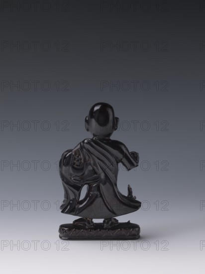 Buddhist monk, Chinese, Qing style, Chinese Buddhism, Qing 1644–1911, Luohan iconography, 19th century, China, sculpture figurine, height 11.8 cm, width 7.1 cm, depth 2.0 cm, A small glossy dark sculpture of a standing figure viewed from behind, rendered in shiny black with smooth reflective surfaces; the figure wears flowing drapery with visible folds and a cloak-like garment, and stands on an oval base that is also dark and polished; the background is a smooth gradient ranging from dark gray at the top to pale gray at the bottom.
