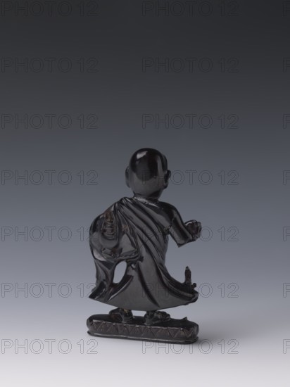 Buddhist monk, Chinese, Qing 1644–1911, 19th century, Luohan iconography, sculpture figurine, height 11.8 cm, width 7.1 cm, depth 2.0 cm., A shiny dark statue viewed from behind showing a robed figure standing on a small decorative base, the figure’s cloak draping with folds and a rounded head with visible ears; the surface is glossy and reflective with visible highlights, and the colors present are black, dark brown, and shades of gray against a gradient gray background.