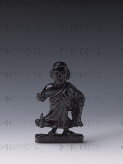 Buddhist monk; unknown creator; Chinese; Qing dynasty 1644–1911, 19th century; Luohan iconography; sculpture, figurine; height 11.8 cm, width 7.1 cm, depth 2.0 cm., A small dark glossy figurine of a standing human figure wearing draped clothing is centered against a smooth gradient background. The figure has a rounded head with defined facial features and ears, and an arm extended slightly forward while the other arm rests by the side. The drapery of the clothing shows carved folds and layered textures, and the figure stands on an oval base with a patterned edge. Visible colors include deep black or very dark brown on the figurine, with subtle highlights of red reflections, and a background gradient of dark gray transitioning to lighter gray toward the bottom.