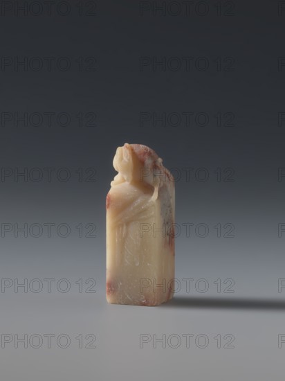 Seal stamp by, late 19th century Qing dynasty 1644–1911, China; carved seal stamp depicting hares; sculpture; height 5.7 cm, width 1.8 cm, thickness 1.7 cm., A small carved object stands upright on a smooth surface against a dark gray to lighter gray gradient background; the object is a rectangular column topped with a sculpted figure reclining over one edge, showing smooth rounded forms and subtle carved folds. The material appears waxy and translucent with a pale creamy color and areas of reddish-brown mottling and streaks; the surface shows fine scratches and slight wear. The overall palette includes shades of cream, beige, reddish-brown, and varying grays in the background and surface, and the object casts a soft shadow to one side.