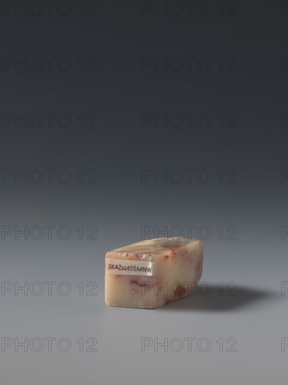 Seal stamp by, gift; depicts hares; Chinese, Qing dynasty 1644–1911, late 19th century; material: sculpture; dimensions: height 5.7 cm, width 1.8 cm, thickness 1.7 cm., A small rectangular block with smooth, slightly mottled surfaces sits on a flat surface casting a soft shadow; the block shows creamy beige and pale yellow tones with reddish-brown streaks and spots, and there is a small pale patch on one end. The background transitions from dark charcoal at the top to mid-gray toward the middle and light gray near the bottom, and the overall scene includes colors of beige, yellow, red-brown, charcoal, and gray.