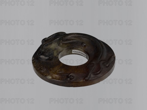 Disc with an image of a lizard;, Chinese, Qing dynasty, Qianlong period 1736–1795. Jade bi disc with stylized antique-style decoration: a lizard, entwines the entire surface. Stone almost black; reverse brown and dark gray-greenish. Type: amulet/ornament. Height 1.6 cm; diameter 8.2 cm., A dark circular carved object with a central hole sits against a pale gray background; the object shows a range of brown tones from dark brown to lighter brown with some greenish-brown areas and darker almost black highlights, and features carved flowing, wave-like raised patterns and a small rounded carved face on one side, with visible surface texture variations and a slightly glossy finish.