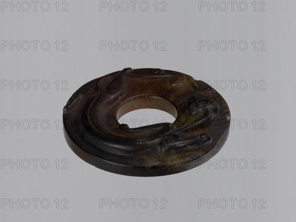 Disc with stylized antique-style decoration showing an entwining image of a lizard, stone almost black with a brown and dark gray-greenish reverse. Amulet/ornament bi disc, dragons iconography. Chinese, Qing dynasty, Qianlong period 1736–1795. Height 1.6 cm; diameter 8.2 cm., A circular carved object with a central hole resting against a light gray background, showing a dark surface with shades of brown, black, olive green, and hints of tan; the carving features smooth, rounded contours and flowing raised forms around the rim, with glossy and matte areas creating subtle highlights and shadows, and the inner edge of the central hole revealing a lighter interior color.