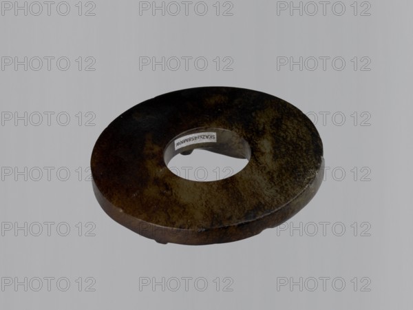 Disc with an image of a lizard, Chinese art, Qing dynasty 1644–1911, Qianlong period/style 1736–1795. Amulet/ornament, bi disc iconography; stylized antique-style decoration with an image of a lizard, entwining the entire surface. Stone nearly black; reverse brown and dark gray-greenish. Date: 1736–1795. Place of origin: China. Varia. Height 1.6 cm; diameter 8.2 cm., A round, flat ring-shaped object with a central circular hole, showing a mottled surface in dark brown and olive green tones, with areas of lighter brown and black; the object casts a faint shadow and rests against a plain light gray background, and a few small surface irregularities and texture variations are visible.