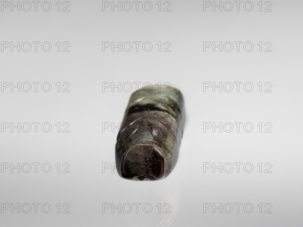 Pig — funerary figurine; creator, Chinese; Han dynasty 202 BCE–220 CE; funerary figurine in jade depicting a pig; animal/zodiac subject; full sculpture; 2nd century BCE–3rd century CE; China; sculpture/figurine; height 2.2 cm; width 9.5 cm; depth 2.4 cm; weight 0.12 kg, A small polished stone with a smooth, rounded rectangular shape rests on a reflective surface; the stone shows a mix of colors including green, dark green, purple, brown, and hints of red, with subtle lighter streaks and darker patches across its surface, and soft reflections beneath it on a pale gray to white background.