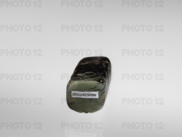 Pig — funerary figurine; creator, Chinese; Han dynasty 202 BCE–220 CE; jade; animal/zodiac subject pig; full sculpture; 2nd century BCE–3rd century CE; sculpture/figurine; height 2.2 cm, width 9.5 cm, depth 2.4 cm; weight 0.12 kg., A small rectangular stone with rounded edges resting on a reflective surface, showing a mottled pattern of dark green, pale green, and black across its top and sides; a small rectangular patch of lighter material is visible on the front face; the background is a smooth gradient of light gray to pale gray.