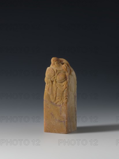Seal stamp, inscription on the stamp; Chinese; Qing dynasty 1644–1911, 18th–19th century; seal stamp; Taoist subject—Immortals, Eight Immortals iconography; sculpture; height 9.3 cm, width 3.2 cm, depth 3.1 cm., A carved stone seal stands on a smooth surface against a dark gray to black gradient background; the stone is primarily warm yellow with subtle areas of brown and faint purple veining, and the carved top features a robed figure with flowing garments and a rounded, smiling face leaning slightly forward while holding a rounded object and a fan, the carving showing detailed folds and rounded contours, and the base of the seal is rectangular with polished, slightly mottled yellow and brown tones.