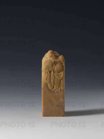 Seal stamp, unknown creator; Chinese; Qing dynasty 1644–1911, 18th–19th century; seal stamp sculpture, Taoist subject depicting the Eight Immortals; inscription on the stamp; materials not specified; dimensions: height 9.3 cm, width 3.2 cm, depth 3.1 cm., A vertical rectangular carved object standing upright on a smooth surface, the material showing warm yellow and brown tones with areas of darker brown and subtle reddish veins; the top portion is sculpted in low relief with a figure wearing flowing robes and holding a fan, the carving exhibiting smooth rounded forms and fine folds, while the lower portion remains mostly plain with natural marbling in brown and reddish hues; the background gradates from a dark charcoal at the top to a lighter gray near the surface, and a soft shadow extends to the right of the object.