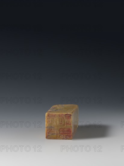 Seal stamp, Chinese art, Chinese culture, Qing dynasty 1644–1911, Qing style, seal stamp, Taoism, Immortals iconography, Eight Immortals, inscription on the stamp, 18th–19th century, China place of origin, sculpture; height 9.3 cm, width 3.2 cm, depth 3.1 cm, The image shows a small rectangular block placed on a flat surface against a dark to light gradient background. The block is primarily yellowish-brown with irregular darker brown streaks and patches. One face of the block displays incised markings filled with a reddish color. The surface of the block appears smooth with slightly rounded edges and minor surface irregularities. The background transitions from dark gray near the top to a lighter gray near the bottom, and a soft shadow in darker gray extends to the right of the block.